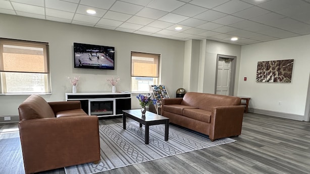 A spacious living room with a brown leather sofa, a coffee table, and a flat-screen TV mounted on the wall. - HomeTowne Studios by Red Roof Richmond, VA - W Broad St (Richmond)
