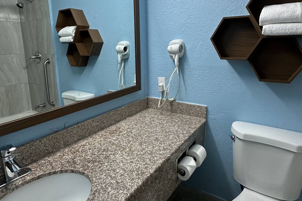 A bathroom with a granite countertop sink, a mirror, two hair dryers mounted on the wall, a toilet, and wooden shelves with towels.