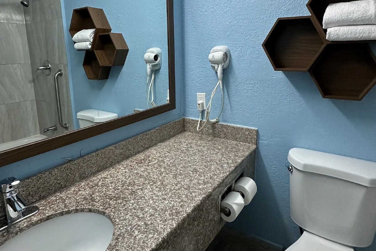 A bathroom with a granite countertop, a white sink, a mirror, two hair dryers, a toilet, and wooden shelves with towels.
