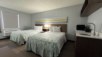 A hotel room with two beds, a large window, a wall-mounted TV, and a kitchenette.