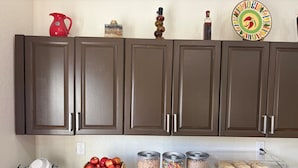 A kitchen with brown cabinets, a fruit basket, cereal containers, and a toaster. - HomeTowne Studios by Red Roof Richmond, VA - W Broad St (Richmond)