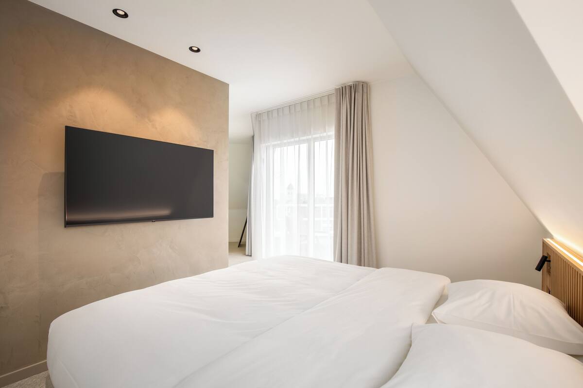 A modern hotel room with a large bed, a flat-screen TV mounted on the wall, and a large window with curtains.
