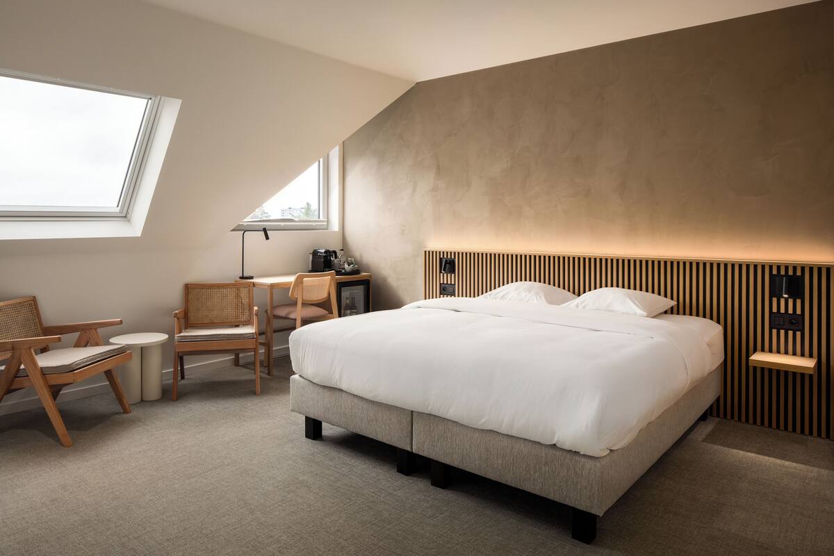 A modern hotel room with a large bed, a desk with a chair, a small table, and a window providing natural light.