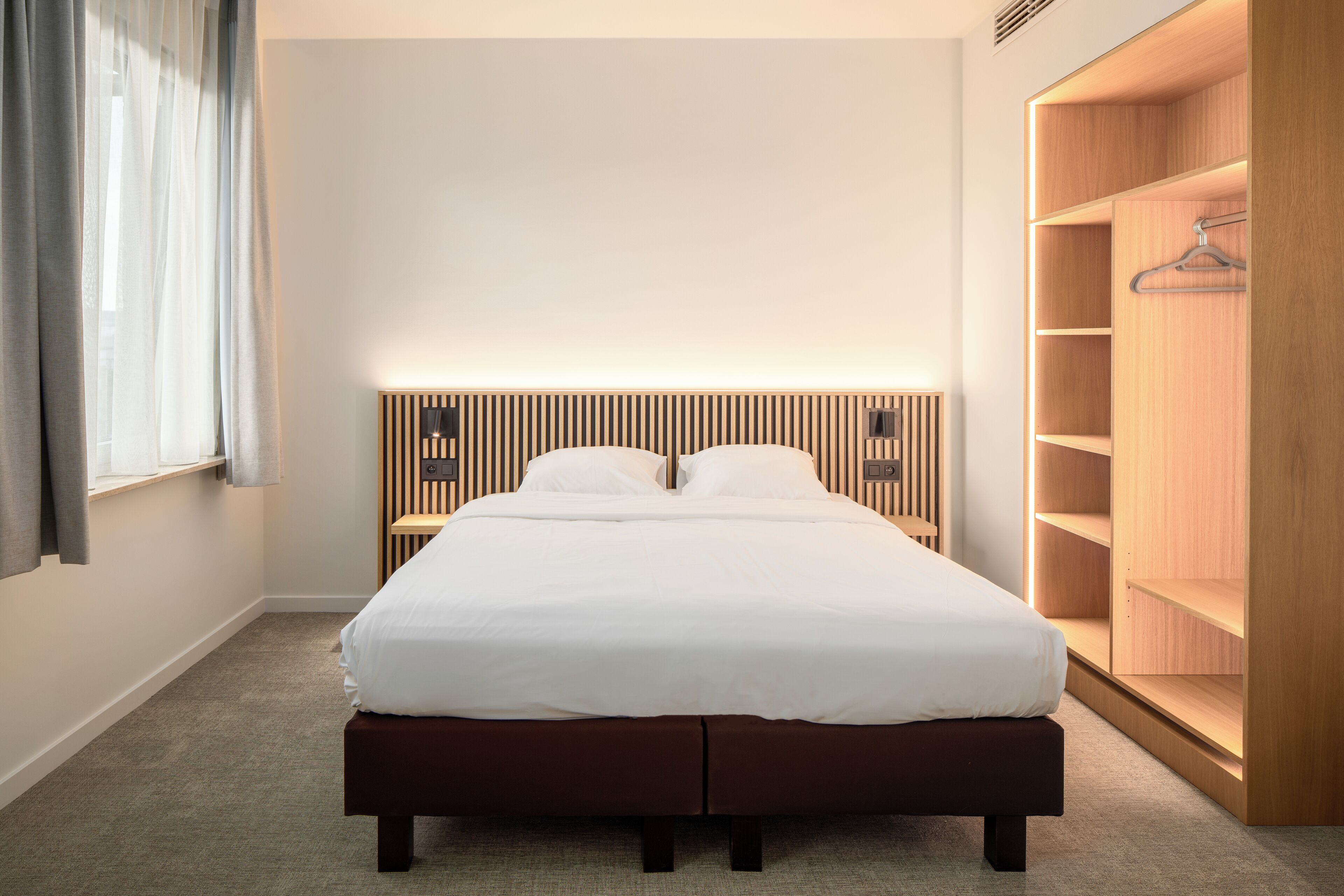 A neatly arranged hotel room with a large bed, a wooden headboard, a bedside table, and a built-in wardrobe.