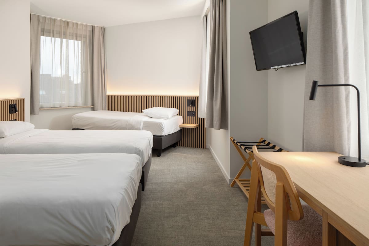 A hotel room with two beds, a desk, a chair, a TV mounted on the wall, and a window with curtains.