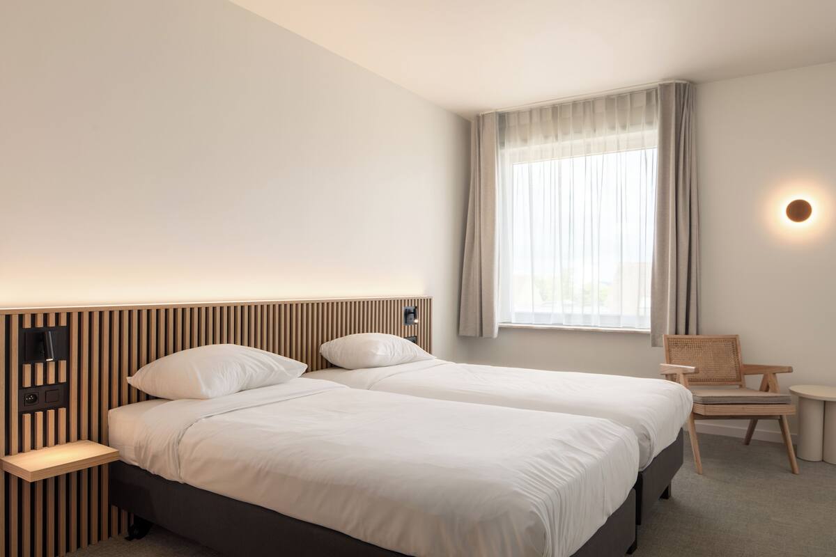 A hotel room with two beds, a nightstand, a chair, a small table, and a window with curtains.