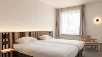 A hotel room with two beds, a nightstand, a chair, a small table, and a window with curtains.
