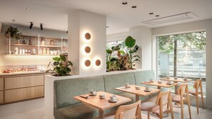 Un café moderne avec des tables et des bancs en bois, des étagères remplies de bocaux et de plantes, et de grandes fenêtres donnant sur la rue.