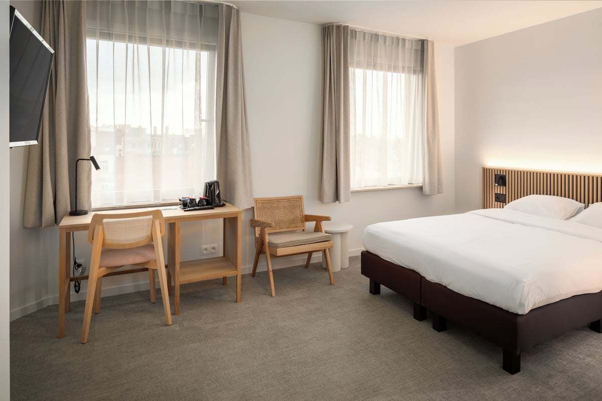 A hotel room with a bed, a desk, two chairs, a TV, and a window with curtains.