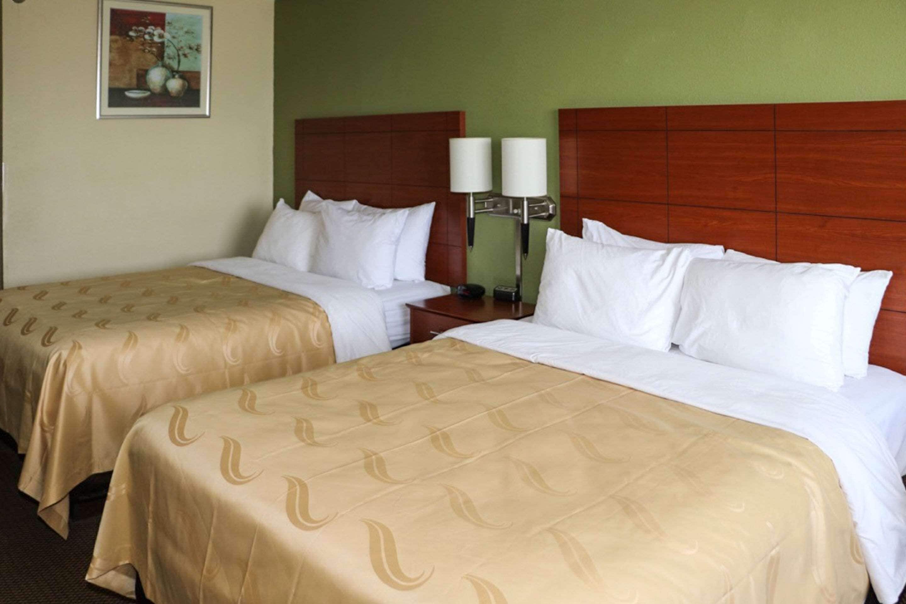 Two single beds with beige bedspreads, a wooden headboard, a nightstand with two lamps, and a framed picture on the wall.