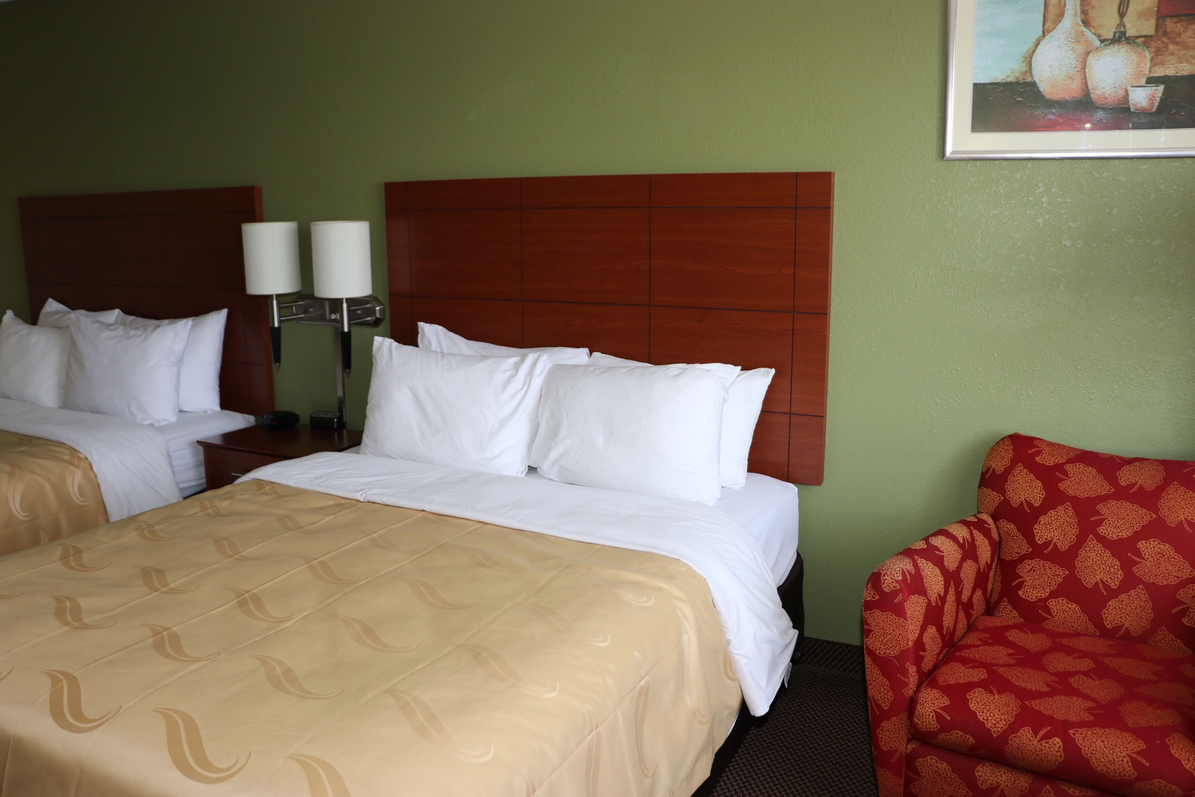 A hotel room with two beds, a wooden headboard, a red patterned armchair, and a painting on the wall.