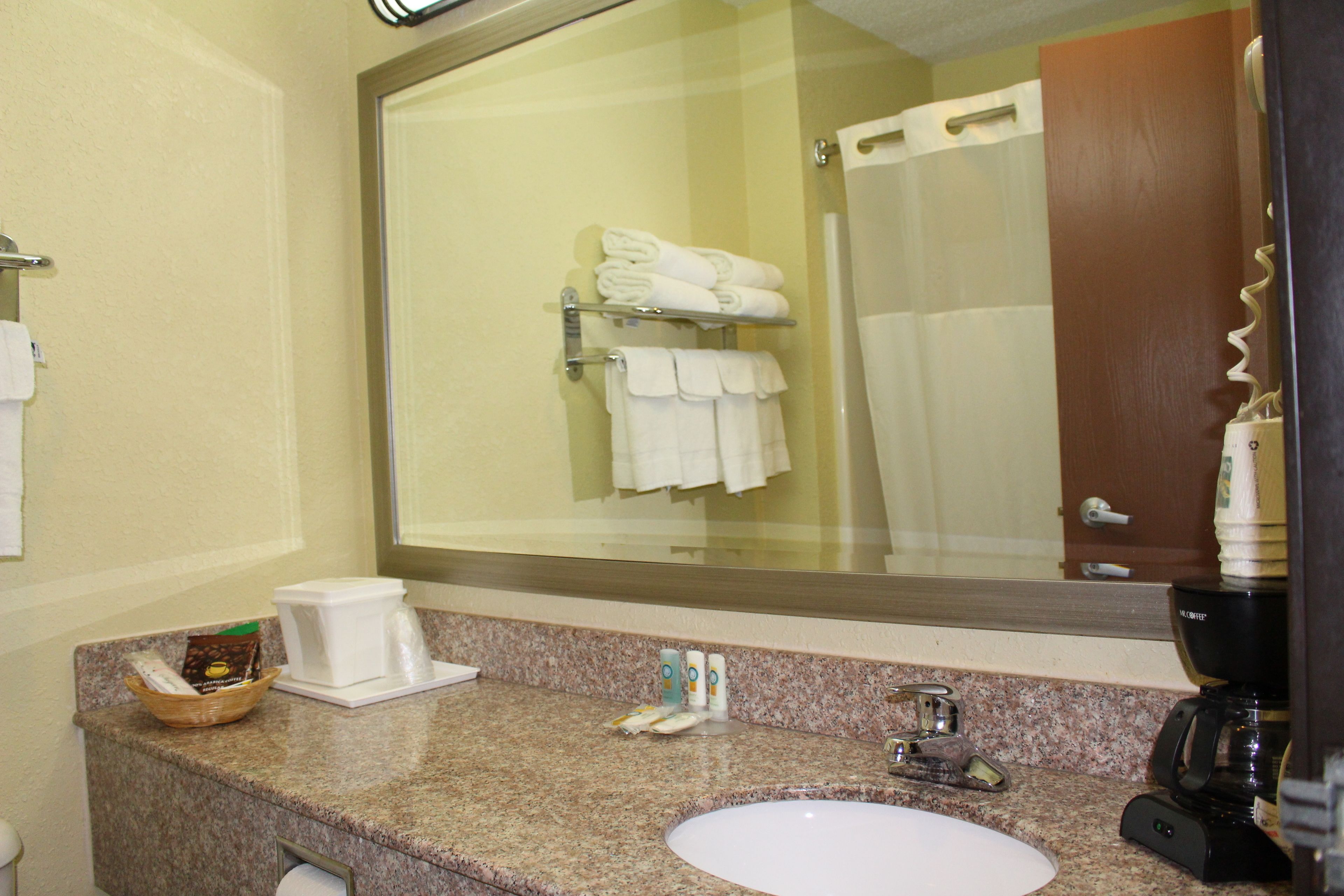 Une salle de bain avec un grand miroir, un lavabo et une machine à café. Des serviettes et des articles de toilette sont posés sur le comptoir.