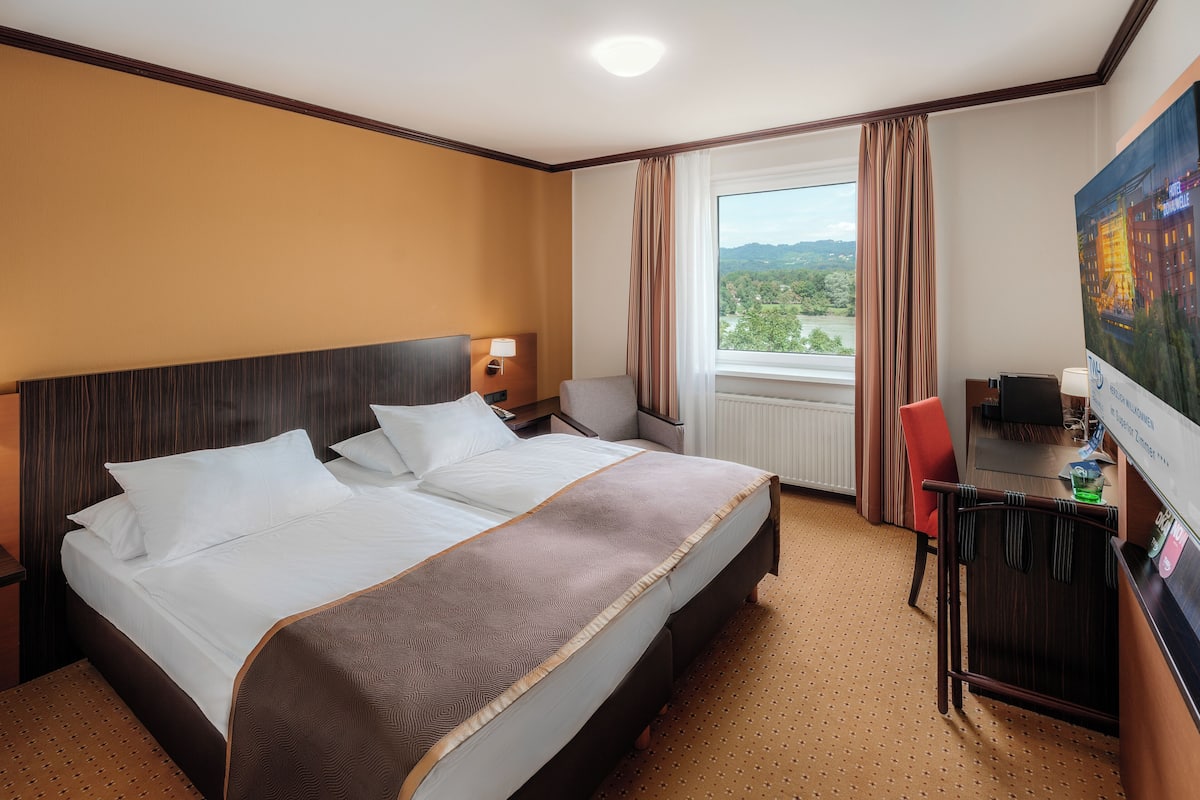 A hotel room with a large bed, a desk, a chair, a TV, and a window with a view of the outdoors.