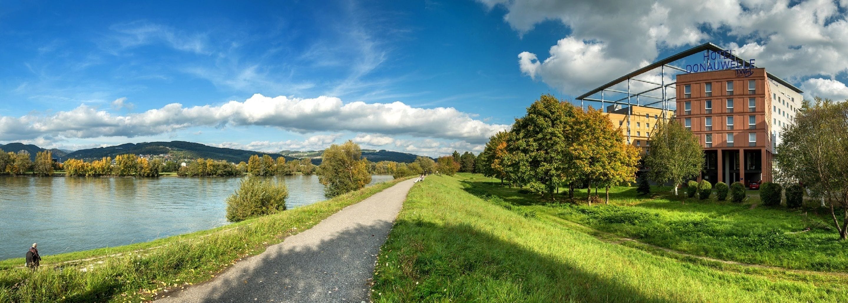 A riverside path leading to a hotel with a modern architectural style, surrounded by lush greenery and a clear sky.