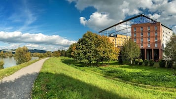 Un sendero a orillas del río que conduce a un hotel de estilo arquitectónico moderno, rodeado de exuberante vegetación y un cielo despejado.