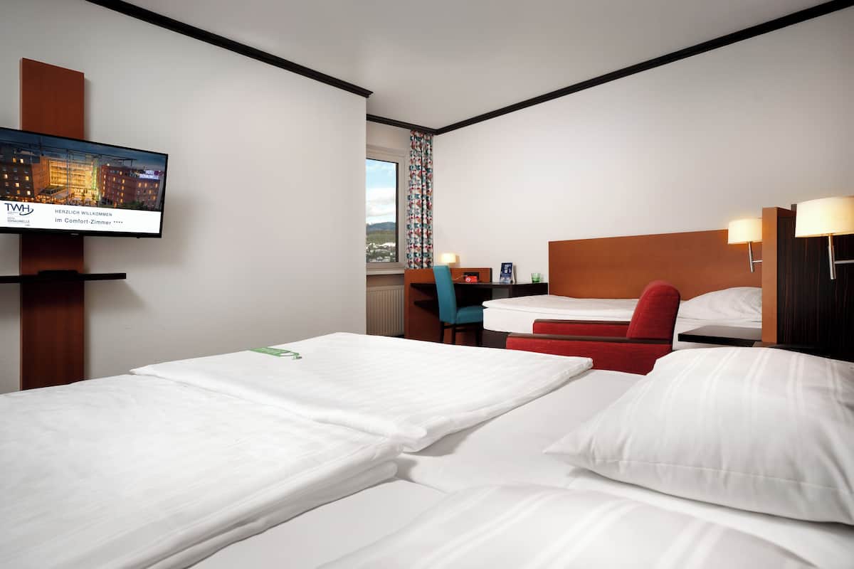 A hotel room with two beds, a TV on the wall, a desk with a chair, and a window with a view.