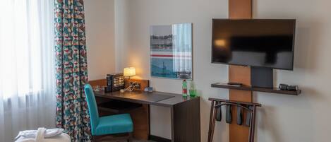 A hotel room with a bed, a desk with a chair, a television, a small table, and a window with curtains.