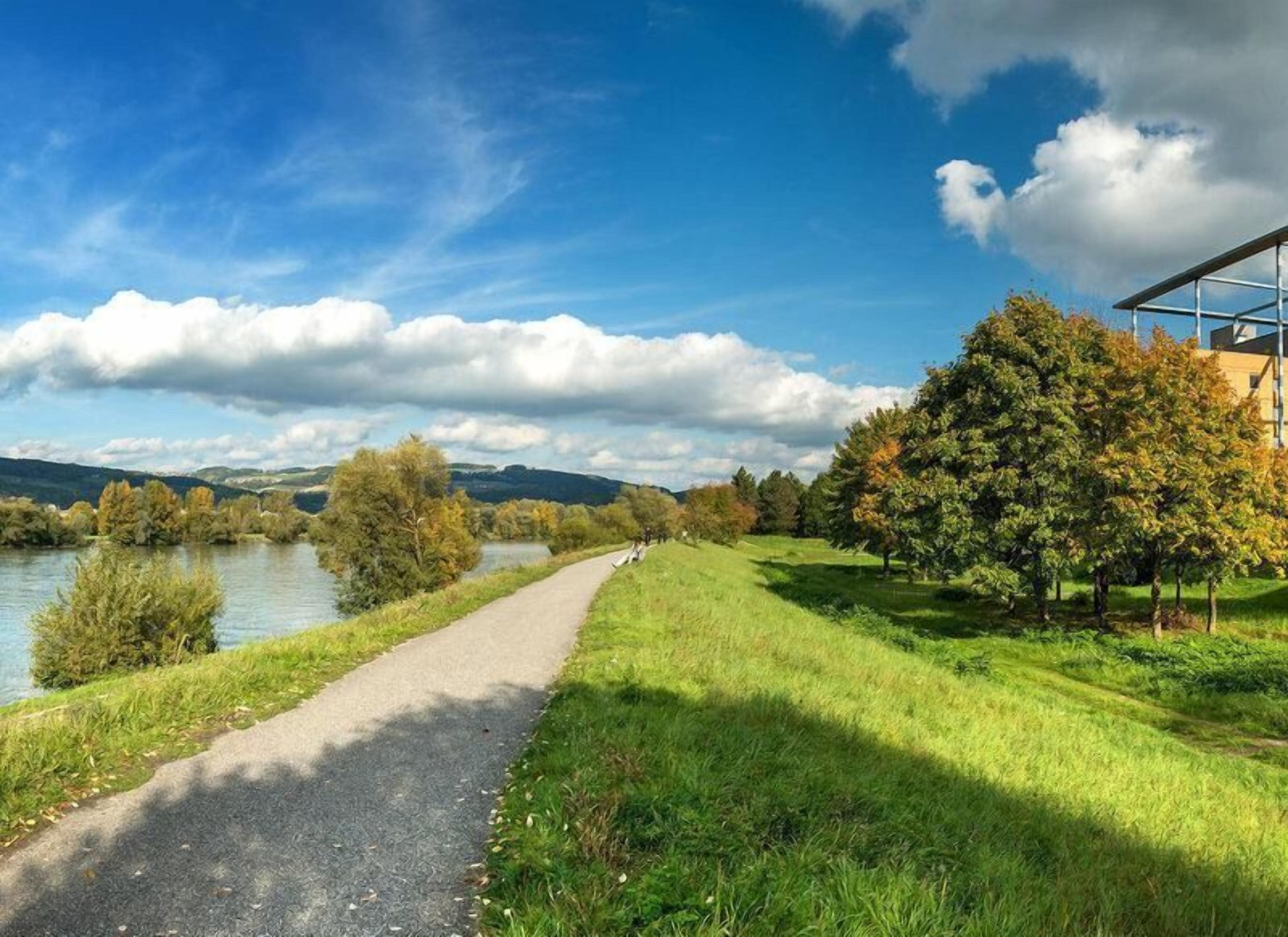 A paved path alongside a river with trees and a building in the background.