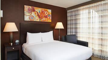 A hotel room with a large bed, two bedside tables with lamps, a chair, a painting on the wall, and a window with curtains.