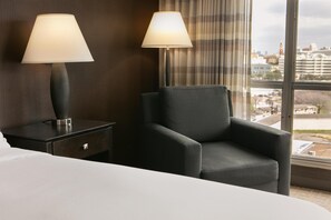 Une chambre d’hôtel avec un lit, deux lampes de chevet, une chaise, une table de chevet et une grande fenêtre donnant sur la ville.