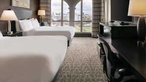 Ein Hotelzimmer mit zwei Betten, einem Schreibtisch mit Fernseher und einem großen Fenster mit Blick auf die Stadt.