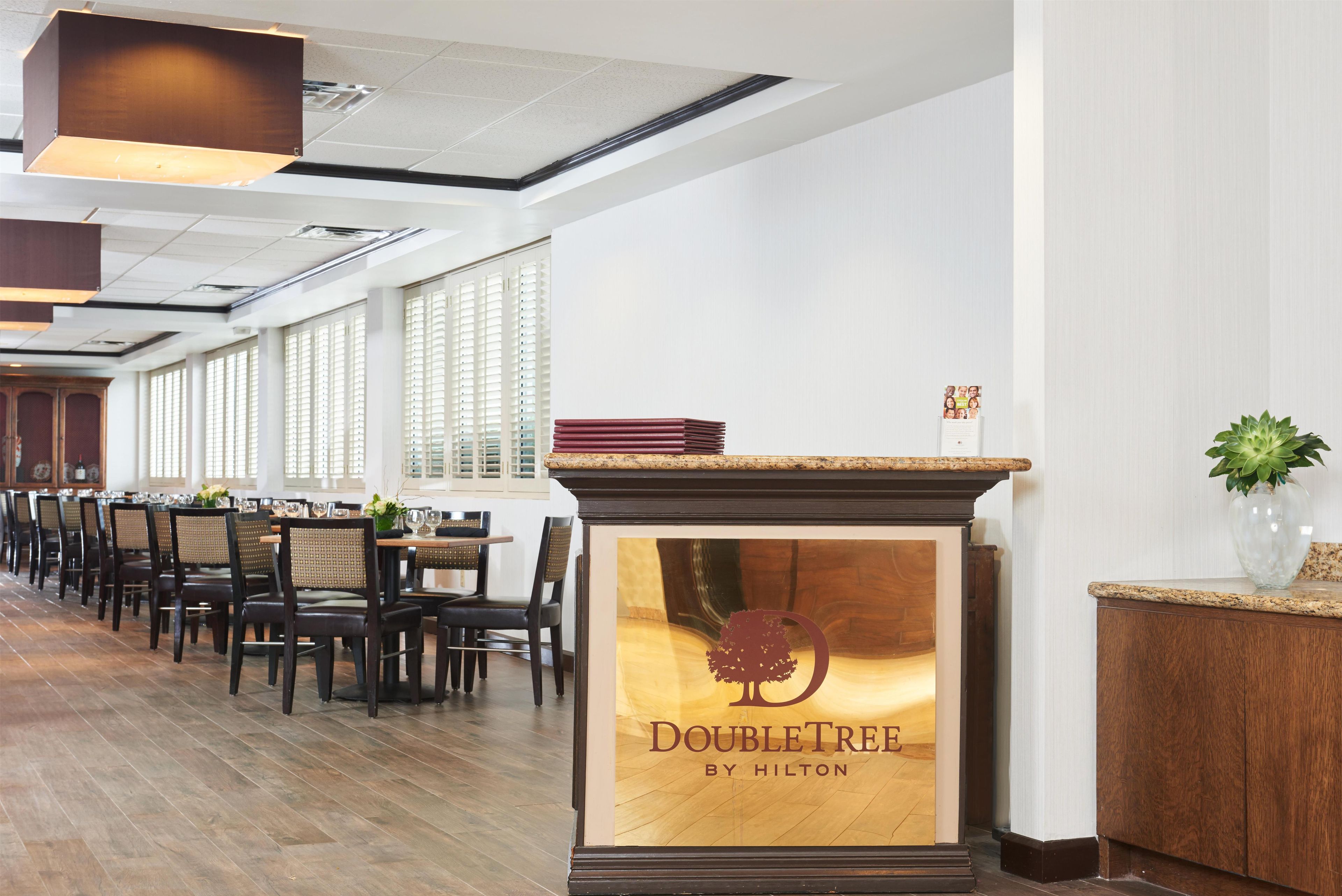 A hotel reception area with a DoubleTree by Hilton sign, wooden flooring, and a long dining area with tables and chairs.