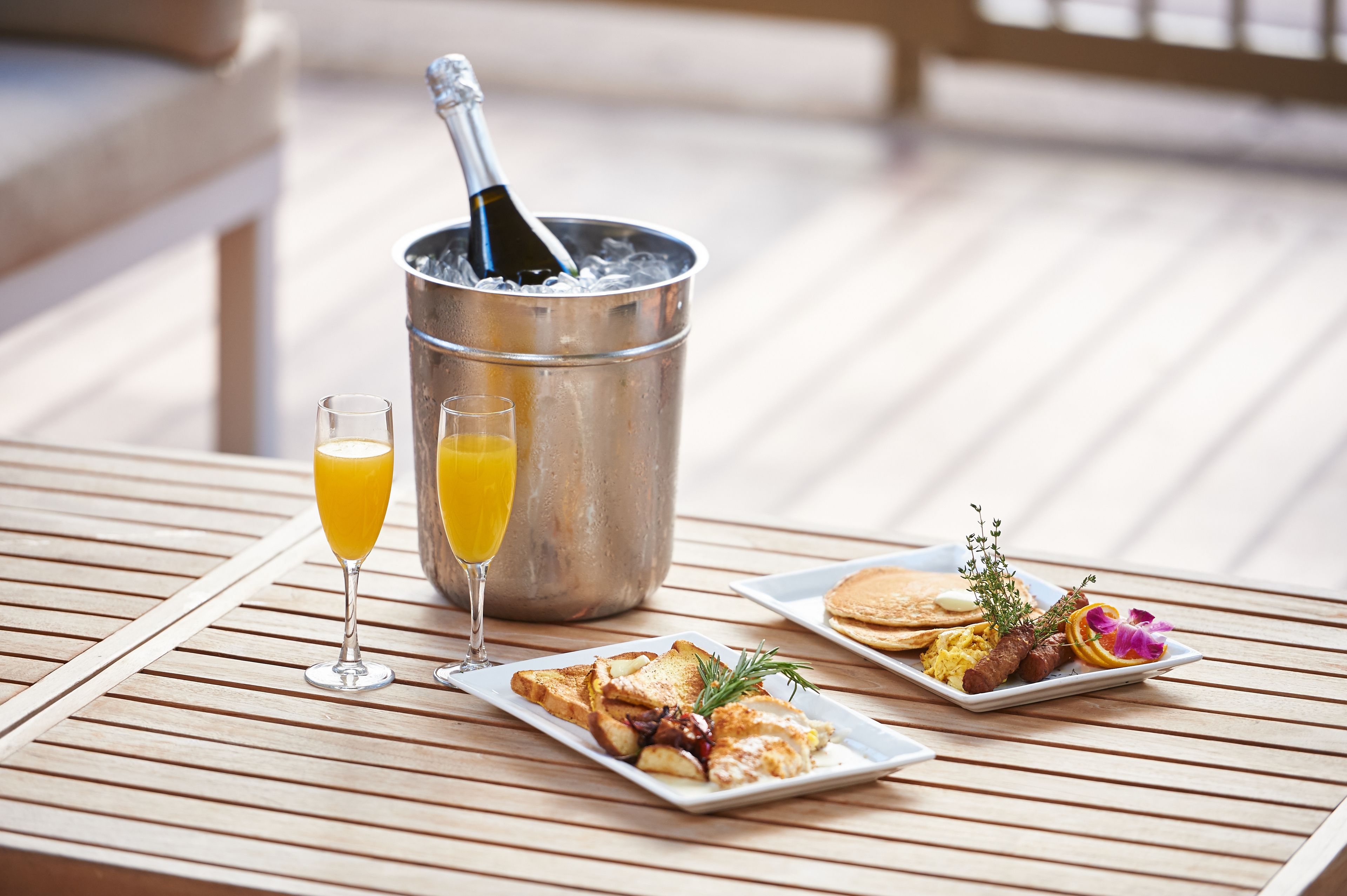 A breakfast setting with a bottle of champagne in a bucket, two glasses of orange juice, and plates of toast, eggs, and pancakes.