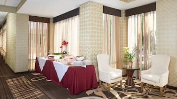 Une chambre d’hôtel avec une table à manger dressée pour un repas, deux fauteuils blancs, une petite table ronde avec une plante et de grandes fenêtres ornées de rideaux légers.