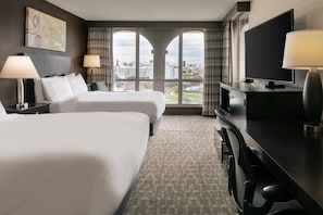 A hotel room with two beds, a desk with a TV, and a large window with a city view. - DoubleTree by Hilton Dallas - Market Center (Dallas)