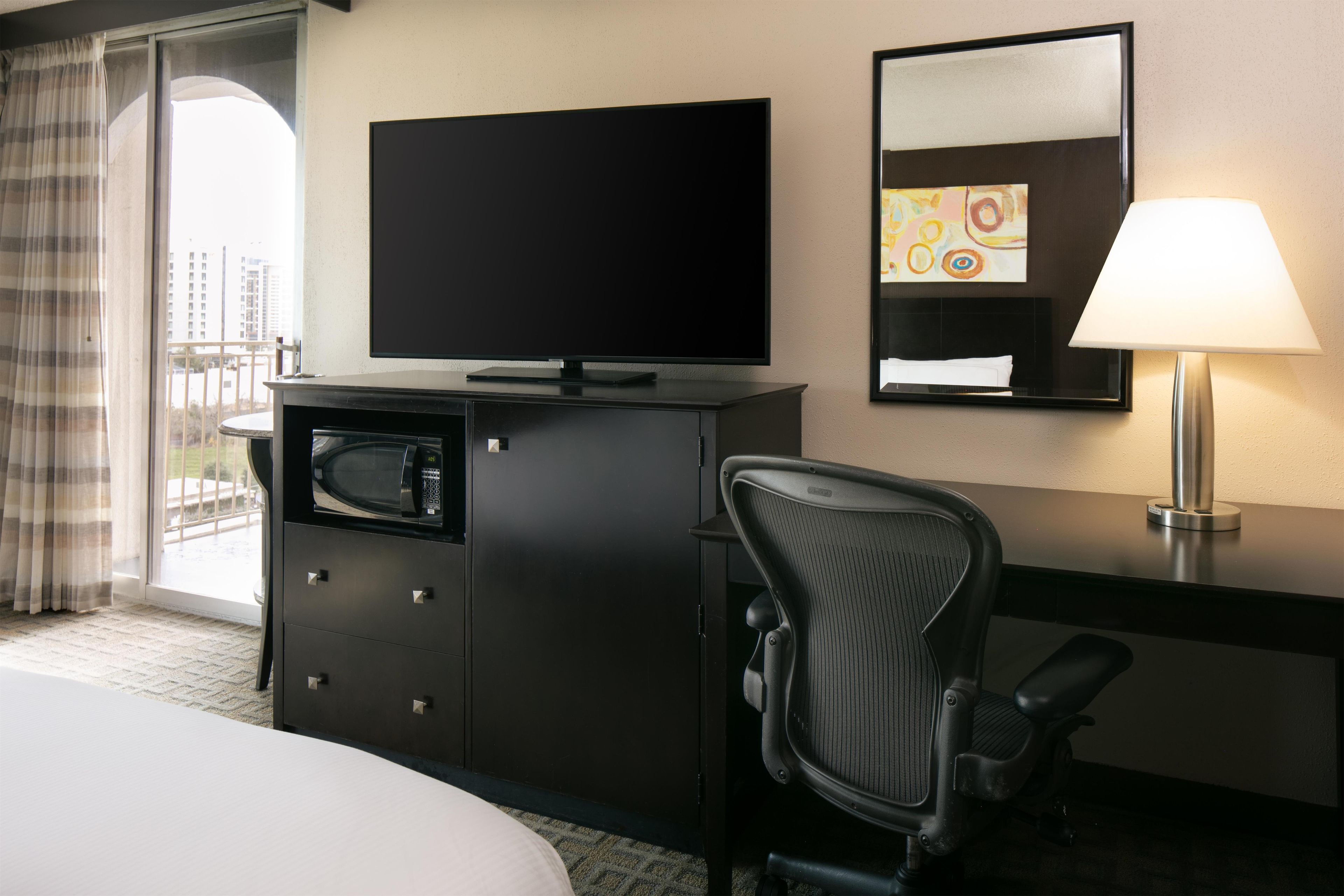 A hotel room with a bed, a desk with a flat-screen TV, a microwave, a chair, a desk lamp, and a window with curtains.