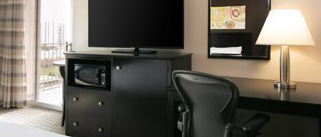 A hotel room with a bed, a desk with a flat-screen TV, a microwave, a chair, a desk lamp, and a window with curtains.