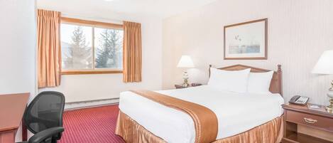 Een hotelkamer met een groot bed, een bureau, een stoel, een raam met gordijnen, een nachtkastje met een lamp en een ingelijst schilderij aan de muur.
