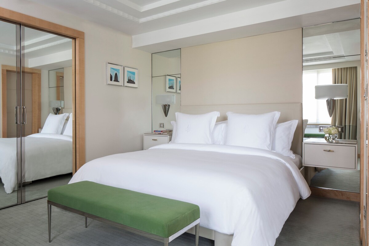 Une chambre d’hôtel avec un grand lit, un banc vert, une table de chevet avec une lampe et un miroir.