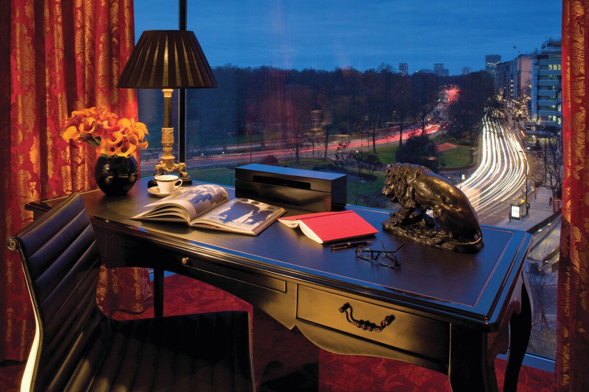 Une chambre d’hôtel avec un bureau, une lampe, un vase de fleurs, un livre, un carnet rouge et une statue.