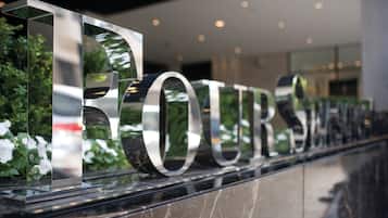 A modern hotel entrance with a reflective sign spelling "HOTEL" and "FOURTH" in large letters, set against a backdrop of greenery and a glass facade.
