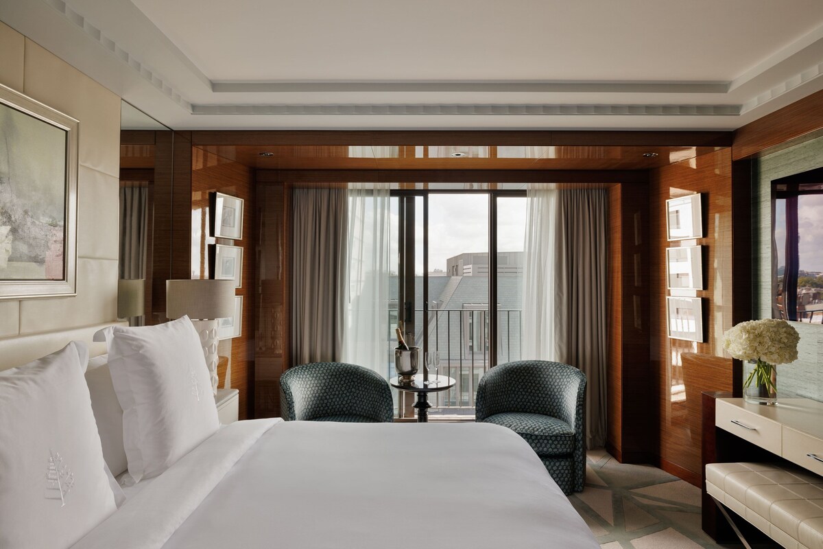 Une chambre d’hôtel avec un grand lit, deux fauteuils, une petite table avec une boisson, et une vue sur les bâtiments à travers de grandes fenêtres.