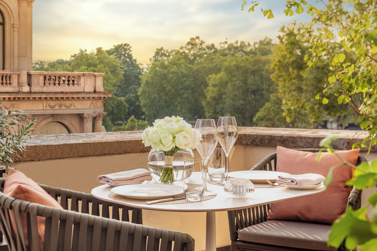 Une table à manger installée à l’extérieur, offrant une vue sur un bâtiment historique et des arbres en arrière-plan.