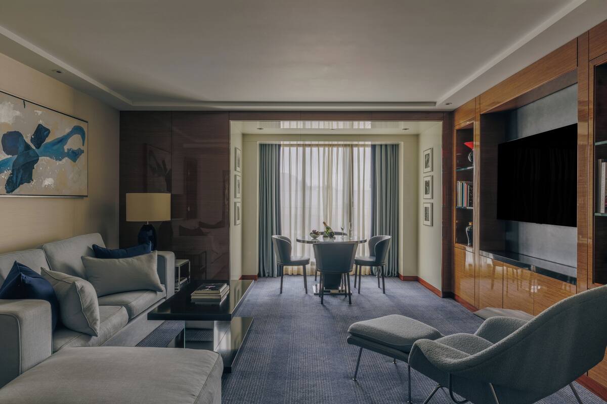 A hotel room with a sofa, armchairs, a dining table with chairs, a TV, and a bookshelf.