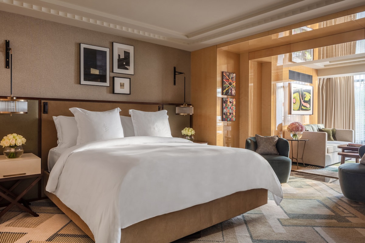 Habitación de hotel con una cama grande, una zona de estar con sofá y sillones, una mesita y obras de arte en las paredes.