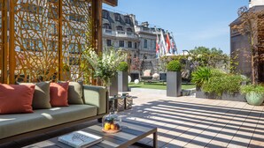 Un espace extérieur moderne aménagé avec un canapé et une table basse, entouré de plantes décoratives et offrant une vue sur un bâtiment historique orné de drapeaux.