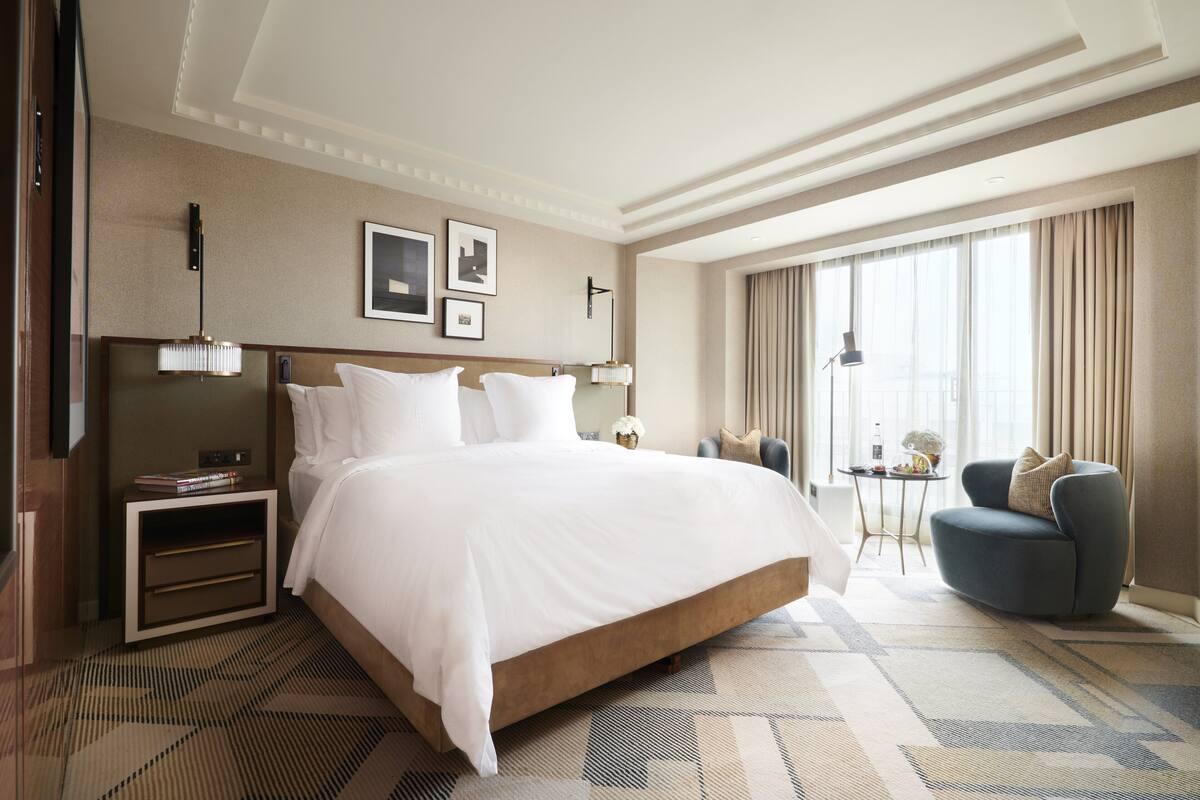 A hotel room with a large bed, a bedside table with a lamp, a small side table, a chair, and a large window with curtains.
