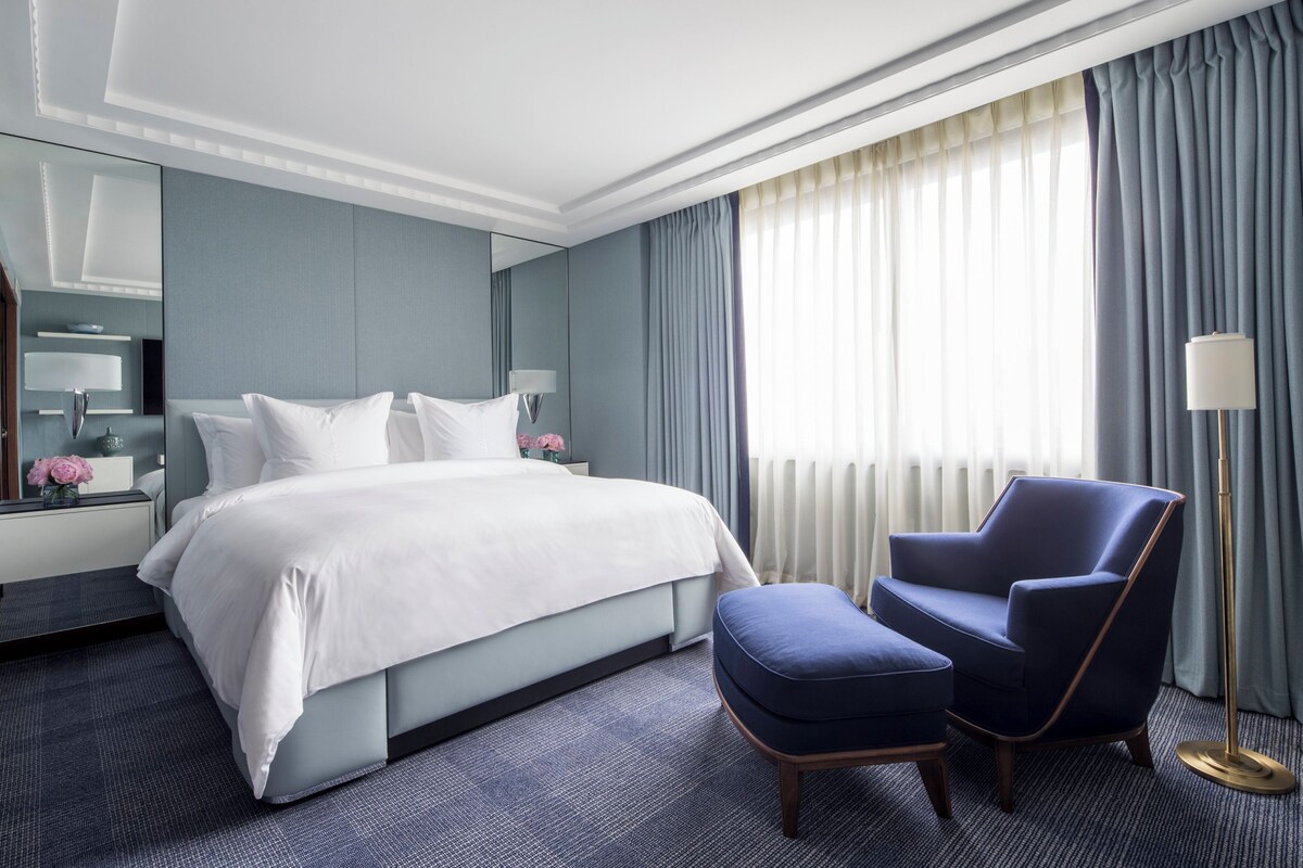 Une chambre d’hôtel moderne avec un grand lit, un fauteuil bleu, un petit pouf, une table de chevet avec une lampe et une grande fenêtre avec des rideaux.