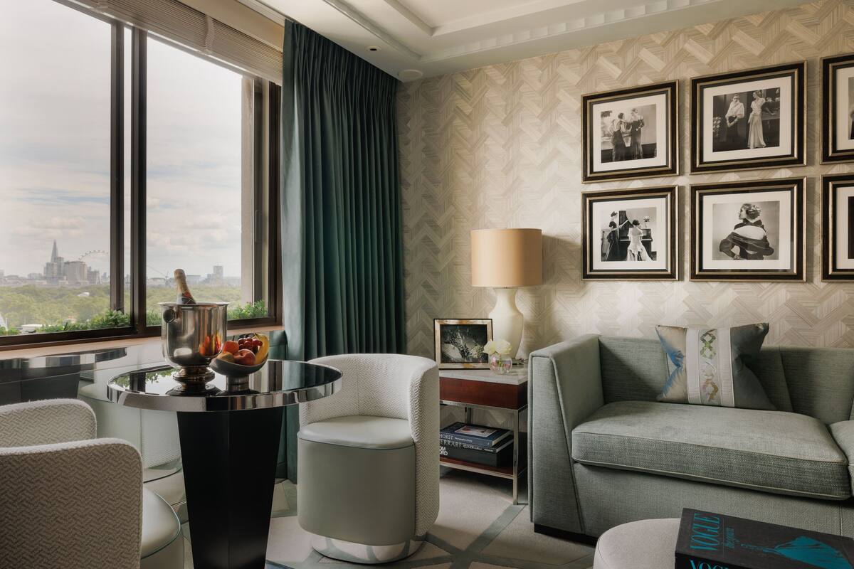 A modern hotel room with a city view, a round table with a fruit bowl, a sofa, and framed photographs on the wall.