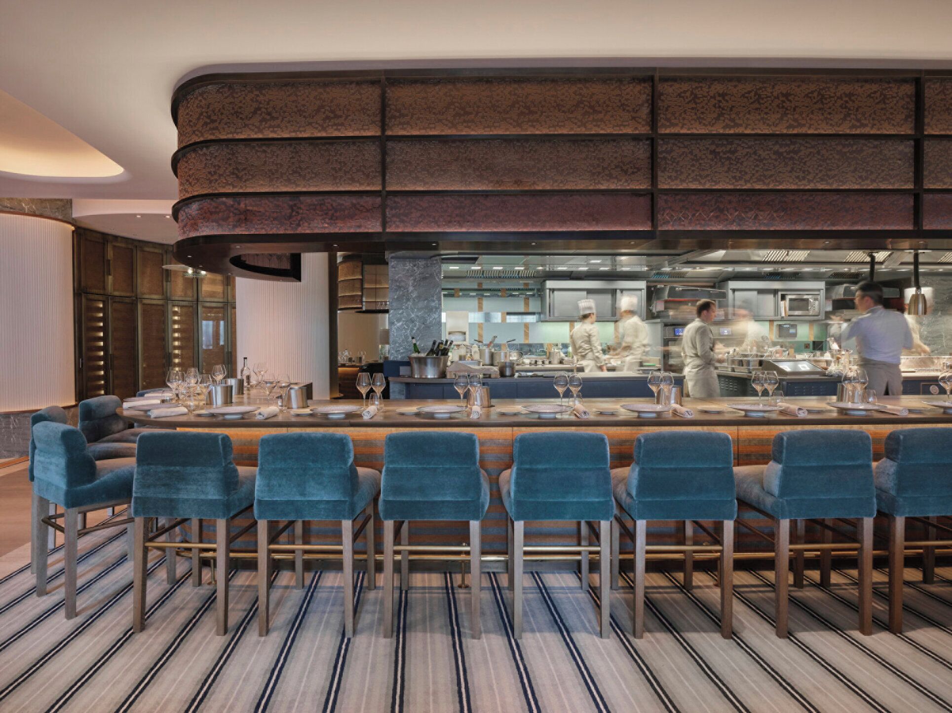 Un restaurante moderno con zona de bar, sillas de terciopelo azul y suelo a rayas.