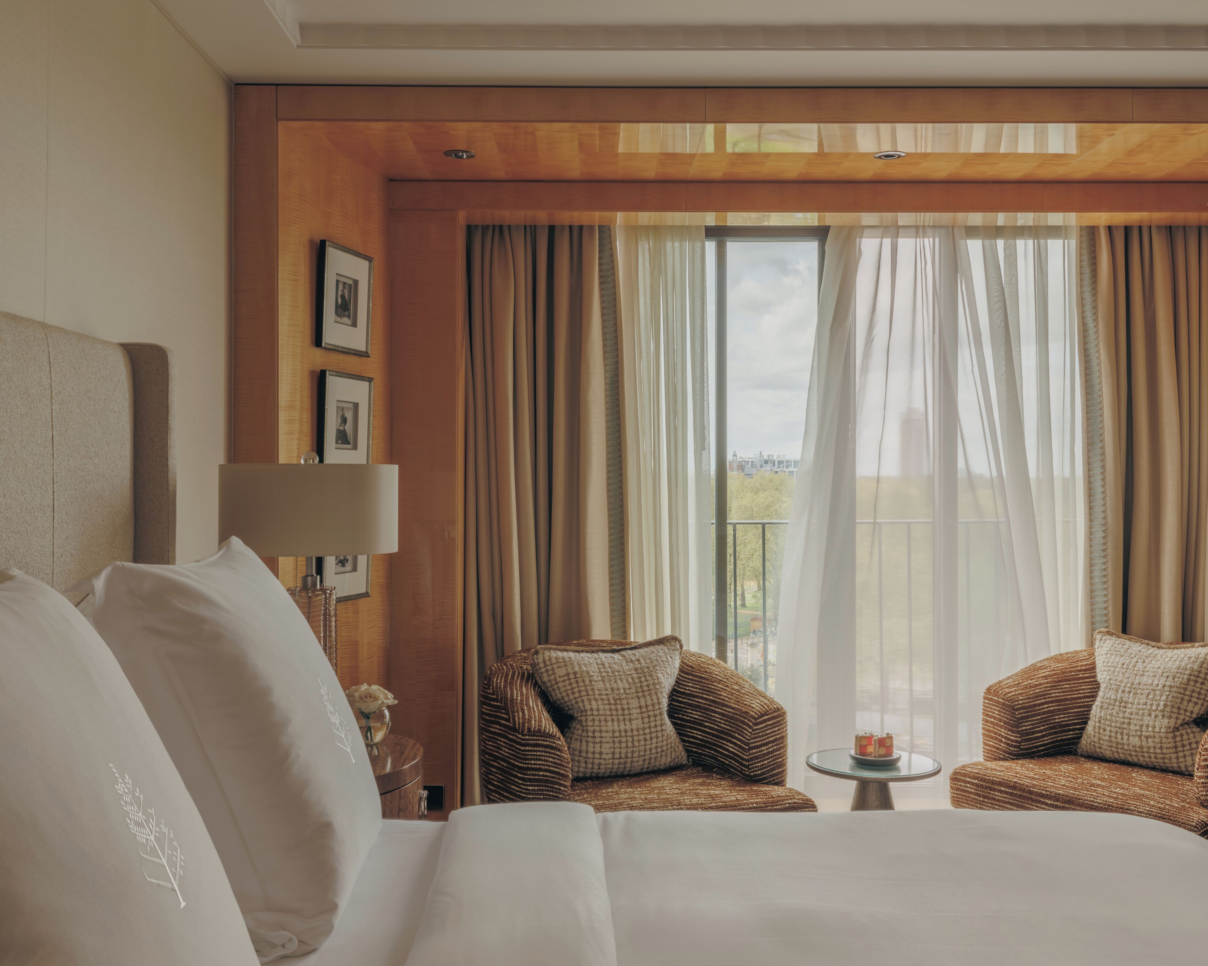 A hotel room with a large bed, two armchairs, a small table, and a view of the city through sheer curtains.