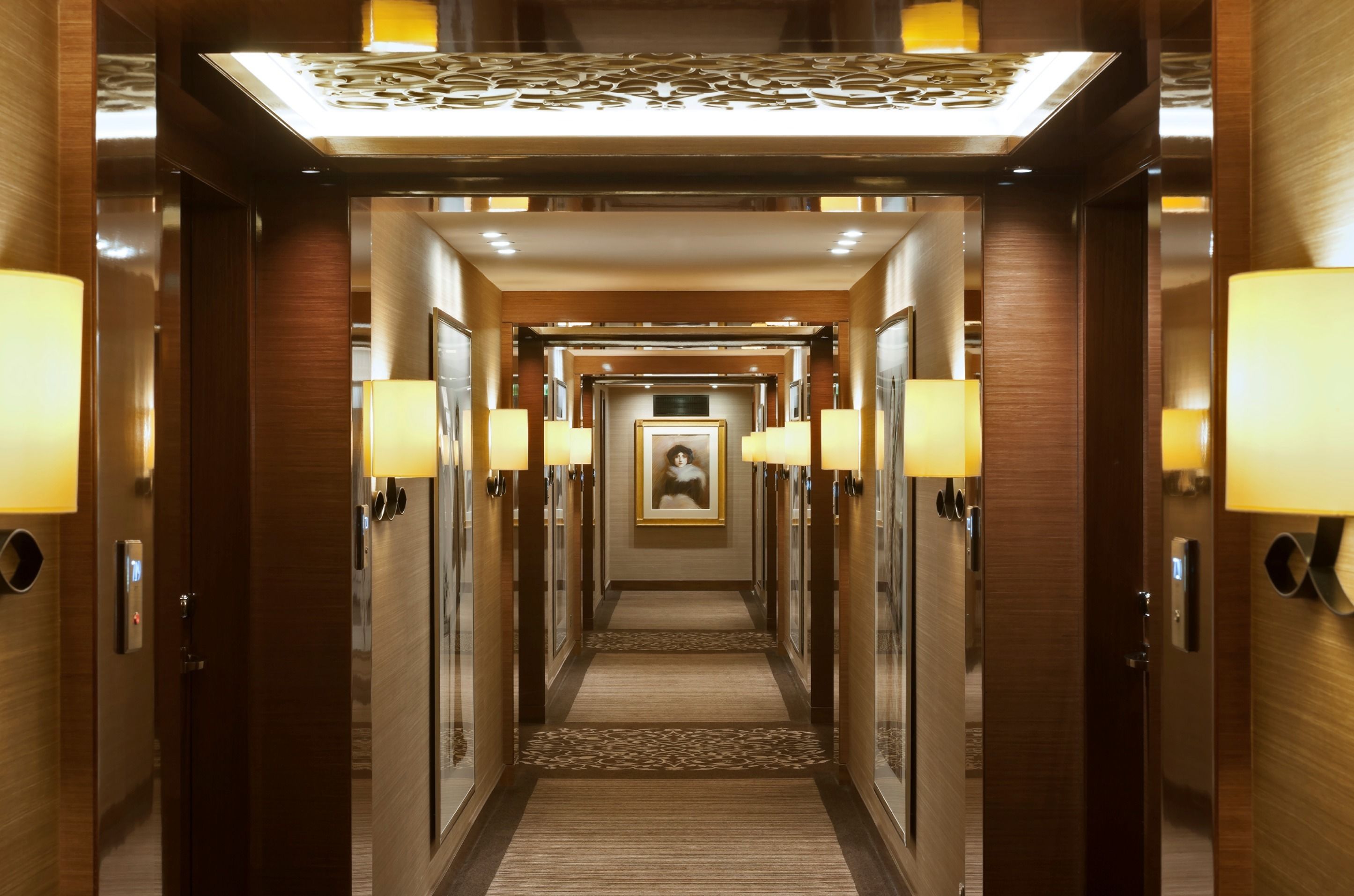 A hotel hallway with a central carpeted walkway, wall-mounted lamps, and a framed picture on the wall.