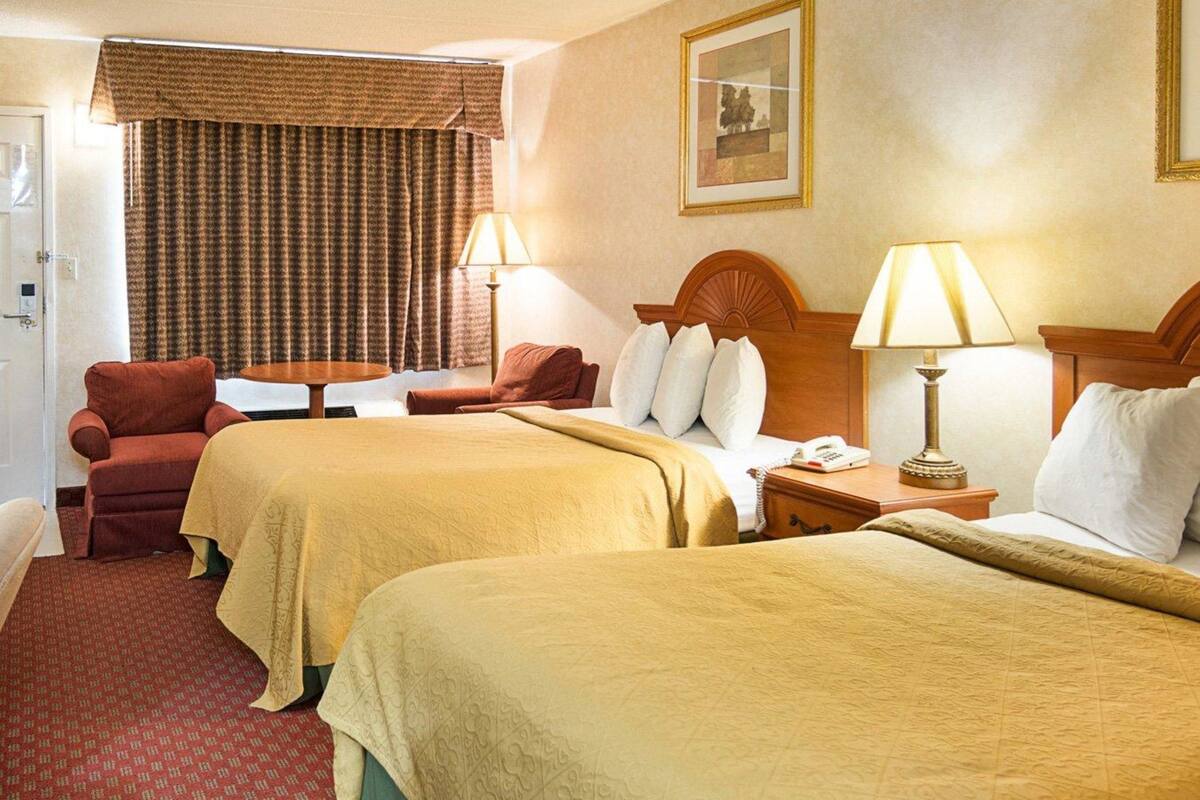 A hotel room with two beds, a desk, a chair, and a lamp.