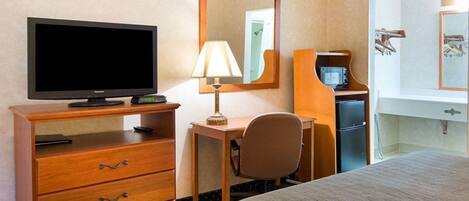 A hotel room with a bed, a desk with a chair, a television, a lamp, a mirror, a refrigerator, and a dresser.