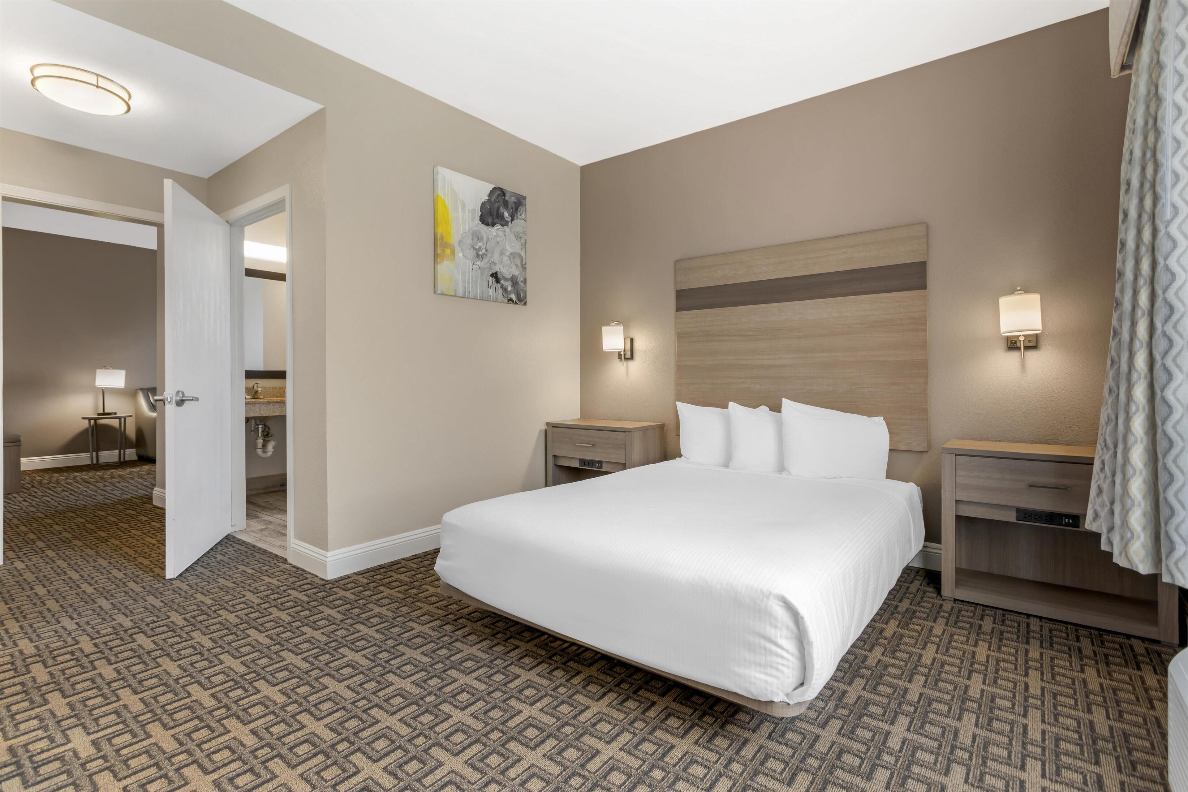 A hotel room with a large bed, two bedside tables, a nightstand, a wall-mounted light, and a door leading to another room.