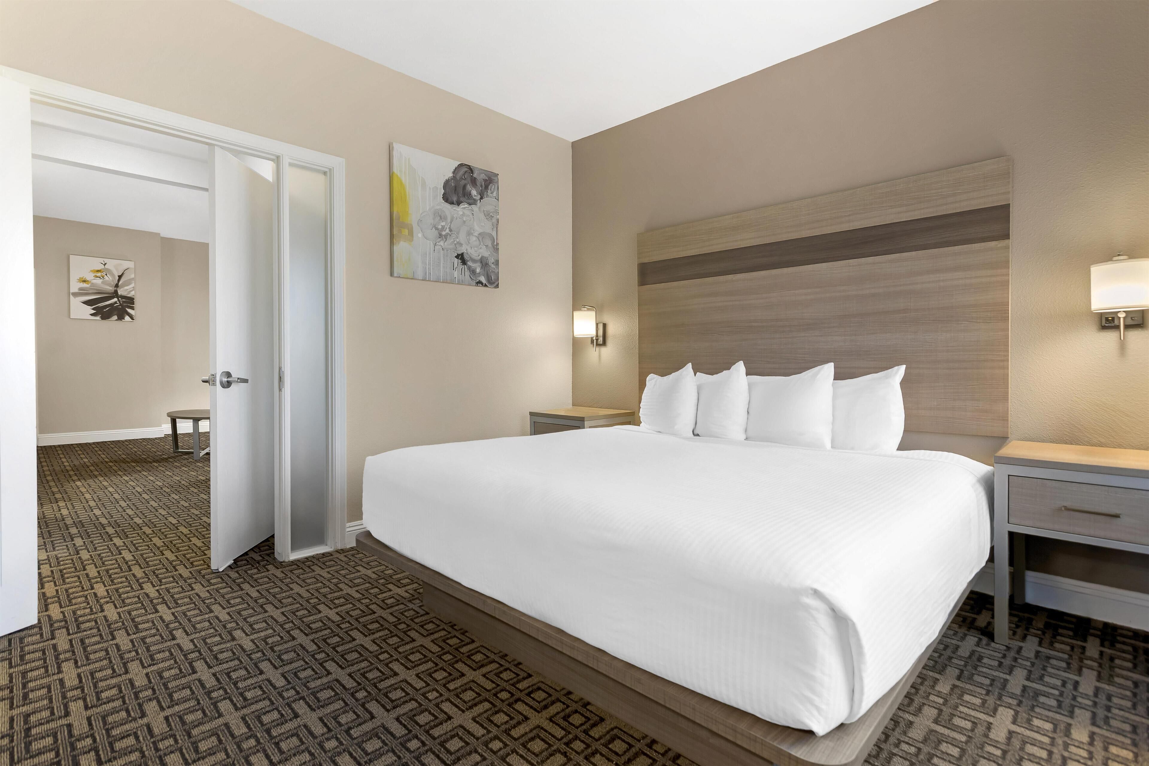 Une chambre d’hôtel avec un grand lit, une table de chevet, une commode, une lampe fixée au mur et une porte donnant sur une autre pièce.